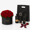 A luxury gift set featuring a round black box of red roses with green leaves, a black gift bag, two black bottles and tubes labeled 'Into the Night', and a dark red ribbon bow on a white background.