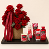 A curated gift set on a black tray featuring a large bouquet of red roses in a woven basket with a red branded ribbon, alongside matching bath and body products including a candle, lotion bottle, hand cream tube, and shower gel. The products and ribbon show the brand name Flowerza and the scent name Japanese Cherry Blossom.