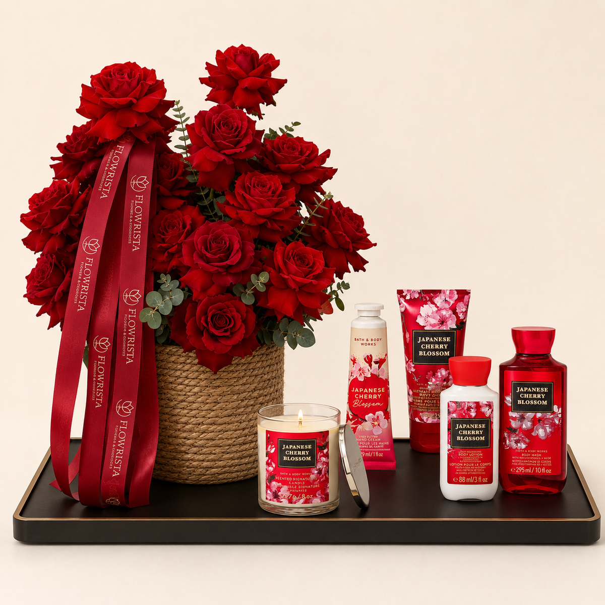 A curated gift set on a black tray featuring a large bouquet of red roses in a woven basket with a red branded ribbon, alongside matching bath and body products including a candle, lotion bottle, hand cream tube, and shower gel. The products and ribbon show the brand name Flowerza and the scent name Japanese Cherry Blossom.