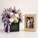 A floral gift box arrangement with purple and white flowers, green foliage, and a lavender ribbon, next to a beige gift bag containing bath and body items. The flower box is labeled FLOWWRISTA FLOWERS & FRAGRANCE.