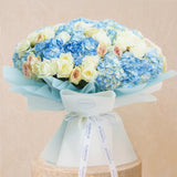 A large floral bouquet featuring light blue hydrangeas, cream and blush pink roses, and pale yellow flowers, wrapped in light blue tissue paper and a white ribbon printed with the brand name 'FLOWERDE'.