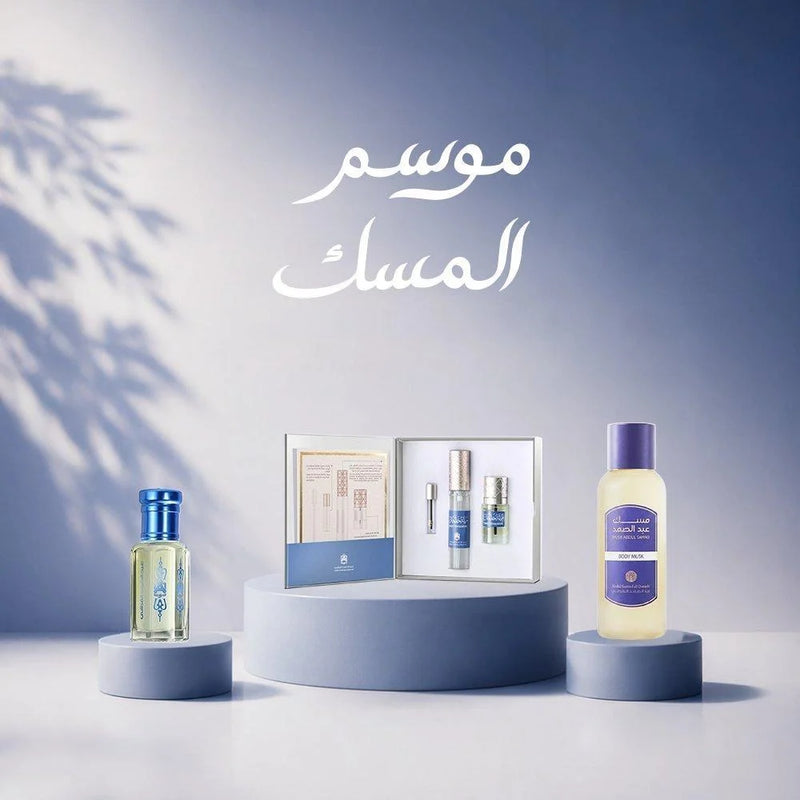 A set of Arabic fragrance and body care products displayed on a minimalist pedestal set against a blue gradient background with Arabic text. The set includes a small clear bottle with a blue cap on the left, an open box with two small blue and silver bottles in the center, and a larger bottle with a purple cap on the right.