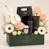 A curated gift basket arrangement from Flowrista featuring a dark green box filled with white and blush peach flowers, greenery, a black tote bag, a black body cream tube labeled Noir, a black body wash bottle, and a small jar of bath salts on a light beige background.