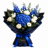 A floral bouquet wrapped in black paper with a dark blue ribbon and a hanging tag reading FLOWWRISTA, featuring large blue hydrangea blooms, white roses, blue delphiniums, and green eucalyptus on a white background.