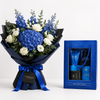 A floral bouquet with blue hydrangeas, blue delphiniums, white roses, and green eucalyptus wrapped in dark blue paper with a blue ribbon, shown beside a blue gift box containing bath and body items labeled Ocean and Flowwrista.
