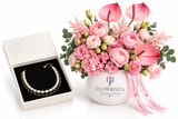 A floral gift arrangement in a white ceramic vase with the brand name FLOWRISTA printed on it, alongside an open cream-colored jewelry box containing a gold and pearl bracelet. The bouquet features pink anthuriums, pink roses, pink carnations, and green foliage, tied with a pink ribbon.
