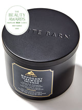 A matte black cylindrical tin candle from White Barn, featuring a gold and black label on the front and 'BARN' embossed on the lid. A white circular 'The Beauty Awards 2023 Best Candle' badge is overlaid in the top-left corner.
