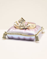 A gold-tone tiara crown with a central clear rhinestone, displayed on top of a small iridescent lavender and pink clutch purse with gold trim and tassels, set against a solid cream background.