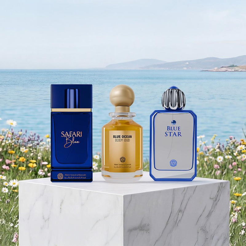 Three perfume bottles from the brand Parfums de Marly are displayed on a white marble pedestal against a scenic coastal background. The bottles are for 'Safari Blue', 'Blue Ocean', and 'Blue Star' scents, featuring deep blue, amber-gold, and light blue designs.