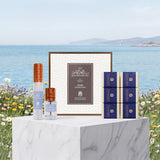 A luxury Arabic fragrance collection displayed on a white marble pedestal with a scenic background of the sea and flowers. The set includes a central framed box, a tall cylindrical bottle with a copper cap, a small spray bottle with a copper cap, and six blue patterned boxes.