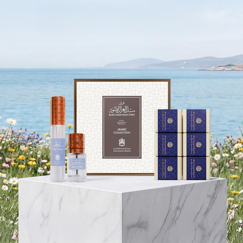A luxury Arabic fragrance collection displayed on a white marble pedestal with a scenic background of the sea and flowers. The set includes a central framed box, a tall cylindrical bottle with a copper cap, a small spray bottle with a copper cap, and six blue patterned boxes.