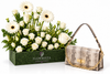 A luxury gift set featuring a green rectangular box of white flowers, including daisies and roses, next to a beige snakeskin-patterned shoulder bag with gold chain and lock details. The box is branded 'FLOWRISTA'.