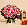 A floral gift set featuring a large bouquet of pink lilies, pink roses, and pink hydrangeas wrapped in black paper with a green ribbon, alongside four Bath Body Works 'Warm Vanilla Sugar' products including a candle, body wash, body lotion, and body spray, arranged on a beige background.
