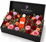 A luxury floral gift box by Flowrista containing a central orange perfume bottle surrounded by an assortment of red, pink, and purple roses with green foliage, presented in an open black box.
