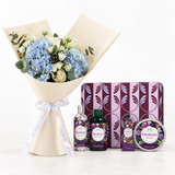 A floral gift set featuring a bouquet of blue hydrangeas, white roses, and green eucalyptus wrapped in beige paper with a white ribbon, alongside a purple patterned box and matching Desuberry body care products including a bottle, tube, and tin.