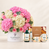 A curated gift set featuring a large round flower arrangement in a white pot with pink and cream hydrangeas, alongside The Body Shop's Almond Milk Collection gift set including an almond milk bottle, lotion tube, soap bar, and tin.