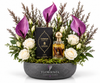 A luxury floral gift arrangement by Flowrista featuring a bottle of Cardamom EDP and its black box nestled in a bowl of white roses, purple calla lilies, green foliage, and moss, set against a solid white background.