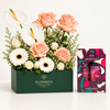A floral gift box arrangement with peach roses, white calla lilies, white gerbera daisies, and baby's breath in a dark green FLOWRISTA box, shown beside a The Body Shop Sugar Plum Duo soap gift set on a clean white background.