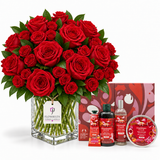 A large bouquet of red roses in a clear glass vase with green stems, displayed with matching red gift items including a box, hand cream tubes, a bottle, and a tin on a white background. The tag on the vase reads Flowrista.