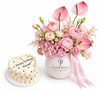 A pink and white flower bouquet in a white ceramic vase with the brand name FLOWRISTA, displayed next to a small heart-shaped birthday cake with white frosting and black text that reads 'Happy Birthday Already'.
