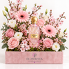A pink diamond-cut perfume bottle with a gold cap nestled in a large floral arrangement of pink roses, pink gerbera daisies, white lisianthus, and pink blossoms, presented in a pink box branded FLOWRISTA.