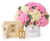 A luxury gift set featuring a large floral arrangement of pink and white flowers in a white bucket labeled 'FLOWRISTA', alongside a beige box, a tube of body lotion, and a small bottle of satin body ood. The products are arranged on a clean white background.