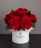A bouquet of vibrant red carnation-like flowers with green leaves arranged in a round white box printed with the brand name 'FLOWRISTA'. The box is placed on a dark surface against a neutral gray background.