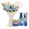 A Valentine's Day gift set featuring a bouquet of light blue hydrangeas, white roses, and eucalyptus wrapped in cream paper with a white ribbon, alongside a blue and gold 'Love is Blue' scented candle, a blue pump bottle of hand and foot wash, and a matching blue tube of body lotion on a white background.