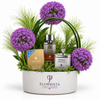 A floral gift basket arrangement in a white oval planter by Flowrista, featuring purple allium flowers, green foliage, and several gift items including a perfume bottle, a candle tin, and a boxed product.