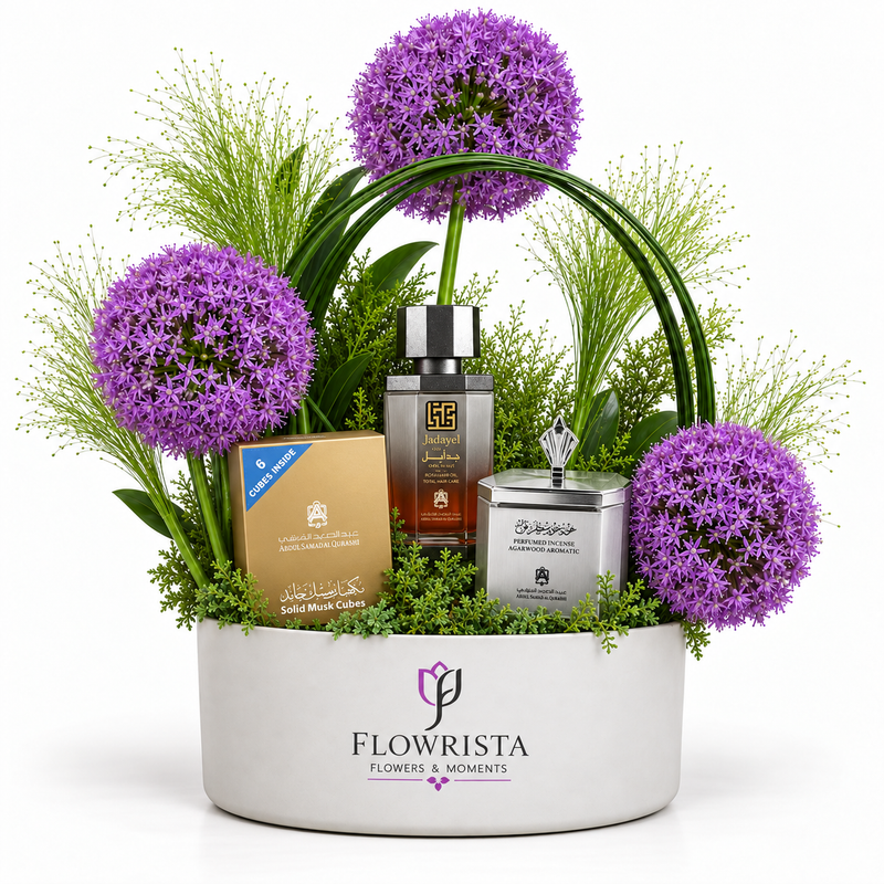 A floral gift basket arrangement in a white oval planter by Flowrista, featuring purple allium flowers, green foliage, and several gift items including a perfume bottle, a candle tin, and a boxed product.
