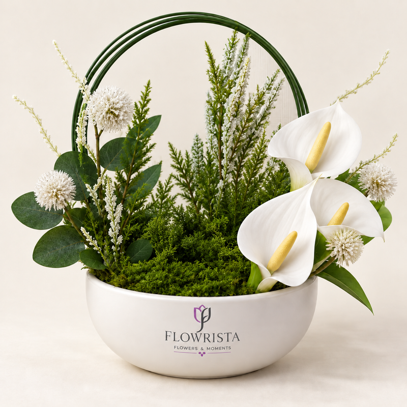 A white ceramic flower basket with a dark green wire handle, filled with green foliage, white calla lilies, and small white clustered blooms. The front of the basket is printed with the brand name 'FLOWRISTA'.