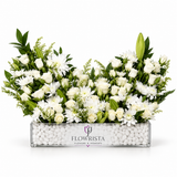 A large floral arrangement of white roses, daisies, and lilies with green foliage, presented in a clear rectangular planter filled with white pebbles. The planter is branded with the logo and name 'FLOWRISTA'.