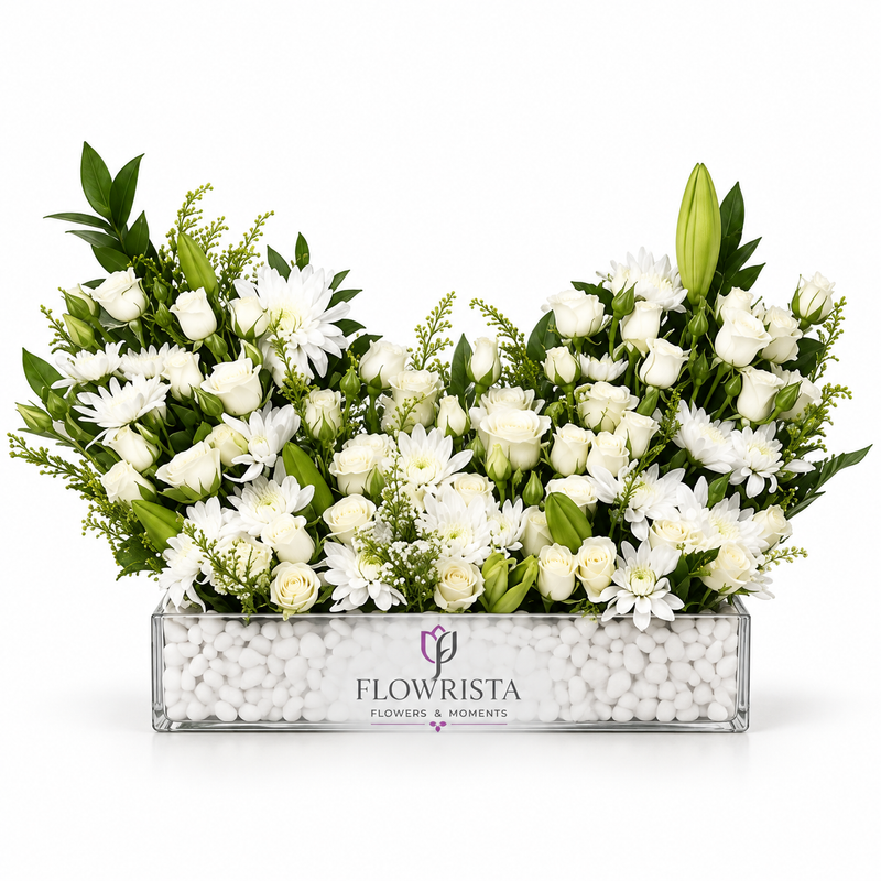 A large floral arrangement of white roses, daisies, and lilies with green foliage, presented in a clear rectangular planter filled with white pebbles. The planter is branded with the logo and name 'FLOWRISTA'.