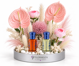 A floral arrangement in a round silver tray with two small bottles of essential oils in front. The arrangement features pink anthuriums, pink roses, pampas grass, and a pink chrysanthemum on white stones.