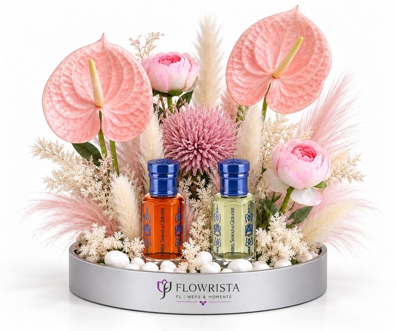 A floral arrangement in a round silver tray with two small bottles of essential oils in front. The arrangement features pink anthuriums, pink roses, pampas grass, and a pink chrysanthemum on white stones.