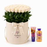 A large round cream gift box filled with white roses and green foliage, tied with a cream satin bow. The box is branded FLOWWRISTA and stands next to two bath/body products from the same brand on a clean white background.