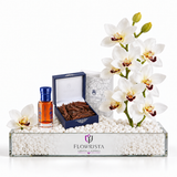 A luxury gift set from Flowrista on a white background, featuring white orchids, a small bottle of amber liquid with a blue cap, a dark blue box of wood chips, and a white candle, all arranged in a clear glass tray filled with white beads. The tray is branded with the Flowrista logo.