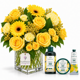 A yellow flower bouquet in a clear square glass vase with a Flowrista tag, alongside three The Body Shop Mango body care items on a white background.