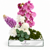 Pink orchids white flowers and purple accents in a mirrored Flowrista tray with white pebbles