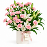 Flowrista pink carnations and white lilies with greenery in a white container tied with a pink ribbon