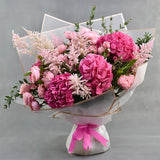 A vibrant bouquet of pink flowers, including hydrangeas, carnations, and pink astilbe, wrapped in white tissue paper with a pink ribbon and clear acrylic stand, set against a neutral gray background.
