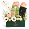 A curated gift set in a dark green rectangular box labeled FLOWWRISTA, filled with white and blush peach flowers, green leaves, and two matching green bottles from Waikiki Body & Beauty. A black notebook with a silver floral design rests on the right side.